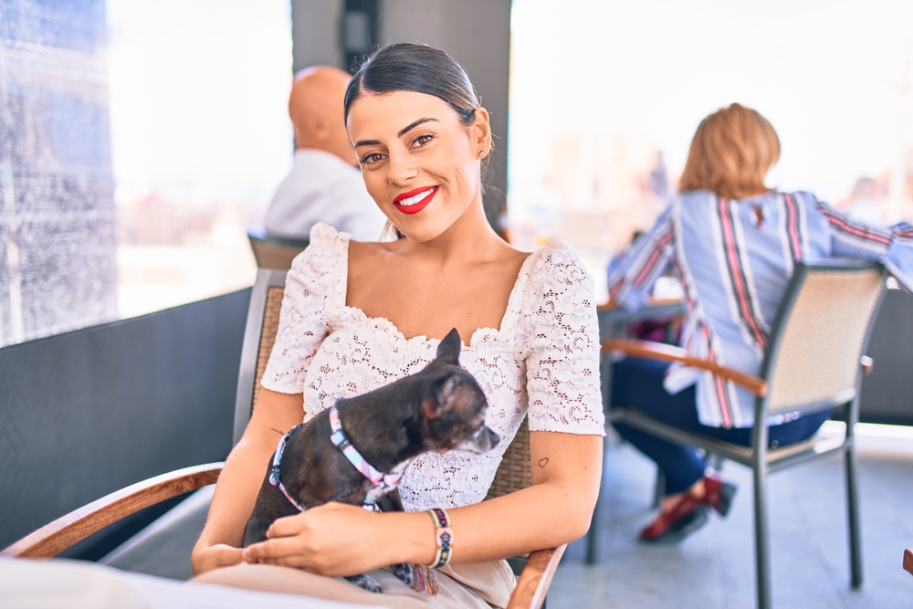 Young brunette woman Sitting with smile on face holding chihuahua dog at restaurant.