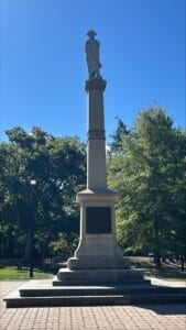 Statue on a tall pedestal in a park setting, surrounded by lush greenery, under a clear blue sky.