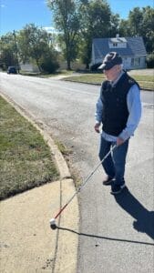 A man, wearing a ball cap walks with a white cane onto a sidewalk on a sunny day.