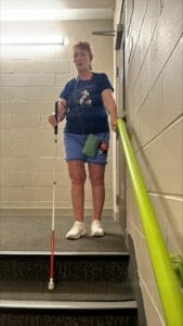 Woman in blue outfit using a white mobility cane and holding onto a green handrail, descending a staircase in a well-lit hallway.