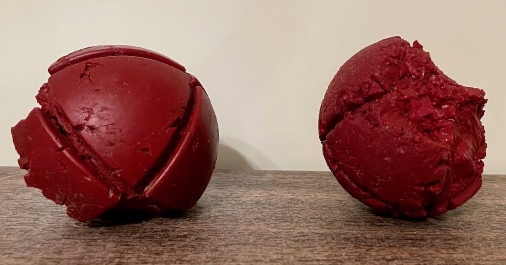 Two old, beat up, chewed red rubber balls on a counter. They have been chewed so much by a dog that almost a third of each ball is gone.