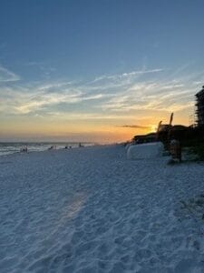 Sunset at a serene beach with soft sand and gentle waves, capturing the peaceful ambiance of the shoreline. Beachgoers can be seen in the distance.