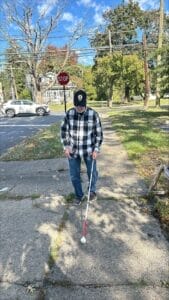 A person with a white cane walking on a sidewalk near a stop sign and a parked car on a sunny day.